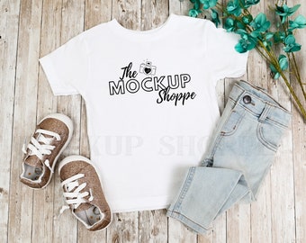 May include: White toddler t-shirt with black text that reads "The Mock Up Shoppe" and a camera graphic with a heart. The shirt is laying on a wooden surface with a pair of light blue jeans and two gold glitter sneakers.