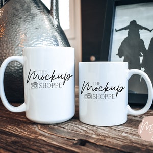 Może przedstawiać: Dwa białe ceramiczne kubki z zakrzywionymi uchwytami są prezentowane na drewnianej powierzchni. Kubki mają napis "THE MOCKUP SHOPPE" w odręcznej czcionce z ikoną aparatu. Dekoracyjny wazon i oprawione zdjęcie znajdują się w tle.