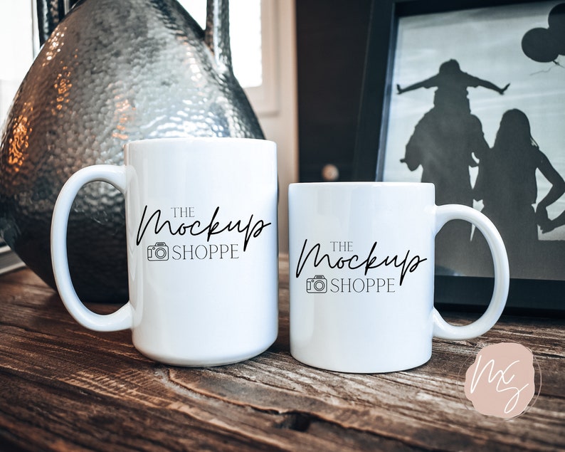 Może przedstawiać: Dwa białe ceramiczne kubki z zakrzywionymi uchwytami są prezentowane na drewnianej powierzchni. Kubki mają napis "THE MOCKUP SHOPPE" w odręcznej czcionce z ikoną aparatu. Dekoracyjny wazon i oprawione zdjęcie znajdują się w tle.