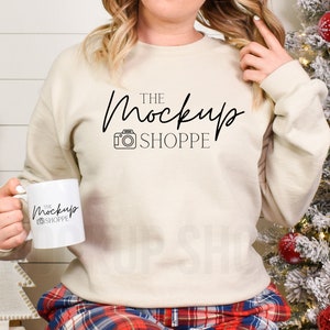 May include: A woman wearing a beige sweatshirt with the text "THE MockUp SHOPPE" and a camera icon. She is holding a white mug with the same text and icon. The background is a white wood panel and a snowy Christmas tree.