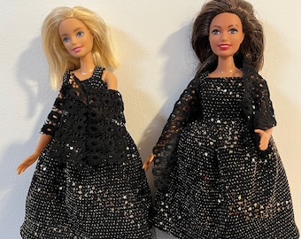 Puede incluir: Dos muñecas con vestidos negros con lentejuelas doradas y encaje. Los vestidos tienen una falda de tul. Las muñecas están de pie sobre una superficie con estampado azul y blanco.