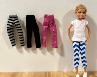 May include: Four pairs of doll-sized leggings in black, pink with a white pattern, and gray and white stripes. A doll wearing a white t-shirt and blue and white chevron leggings.