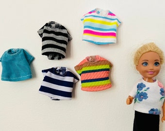Puede incluir: Cinco camisas a rayas en miniatura de varios colores, incluyendo azul, rosa, negro y blanco, y una muñeca que lleva una camisa blanca con un estampado floral.