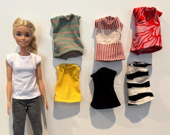 May include: A collection of seven sleeveless tops for dolls, including striped, floral, and solid colors. The tops are made of fabric and are in various colors, including red, white, black, yellow, and green.