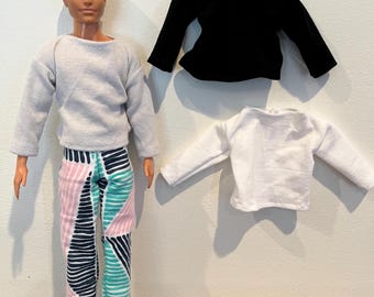 Puede incluir: Una muñeca con un suéter gris claro y pantalones estampados con formas geométricas rosas, turquesas y azul marino. También hay dos suéteres de muñeca adicionales, uno negro y otro blanco. La muñeca lleva zapatos blancos.