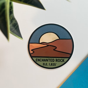 Puede incluir: Una pegatina circular con un dise&ntilde;o de paisaje que representa una puesta de sol sobre un paisaje des&eacute;rtico. La pegatina tiene un borde azul y el texto "ENCHANTED ROCK ELE. 1,825'". La paleta de colores incluye tonos marrones, naranjas y azules.