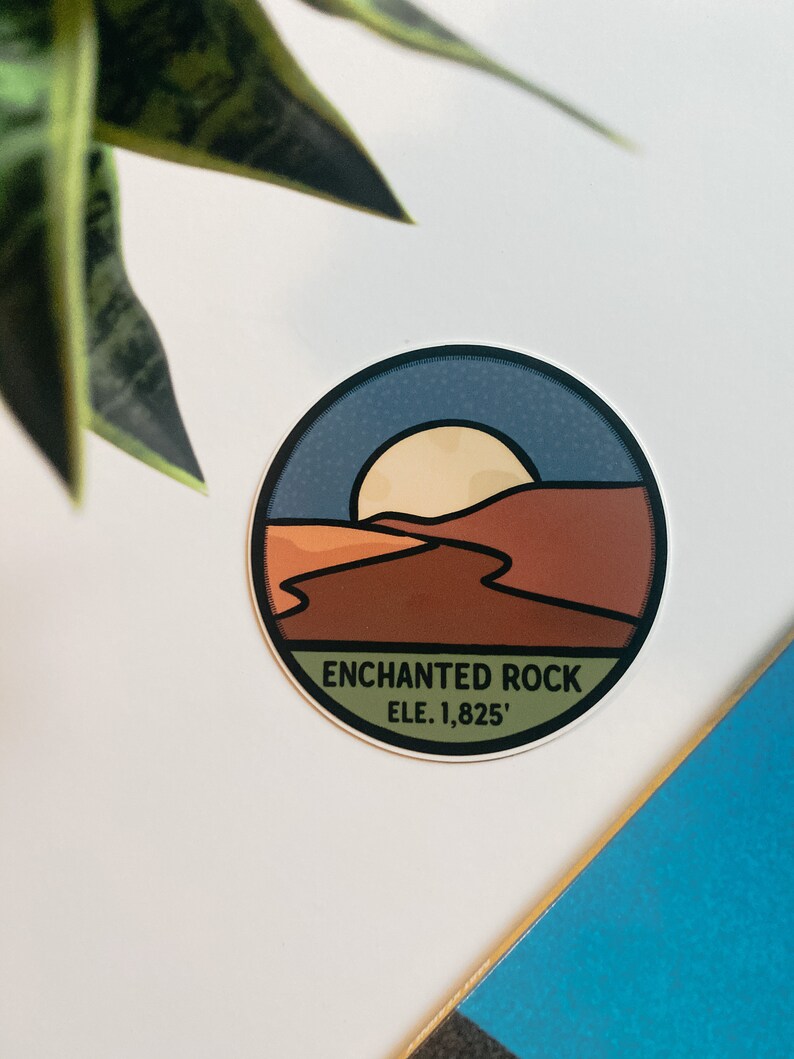 Puede incluir: Una pegatina circular con un dise&ntilde;o de paisaje que representa una puesta de sol sobre un paisaje des&eacute;rtico. La pegatina tiene un borde azul y el texto "ENCHANTED ROCK ELE. 1,825'". La paleta de colores incluye tonos marrones, naranjas y azules.