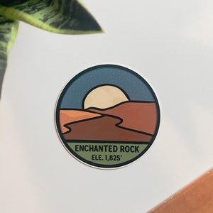 Puede incluir: Una pegatina redonda con un dise&ntilde;o de paisaje que representa una puesta de sol sobre un paisaje des&eacute;rtico. La pegatina tiene un borde azul y el texto "ENCHANTED ROCK ELE. 1,825'". La pegatina est&aacute; sobre una superficie blanca.