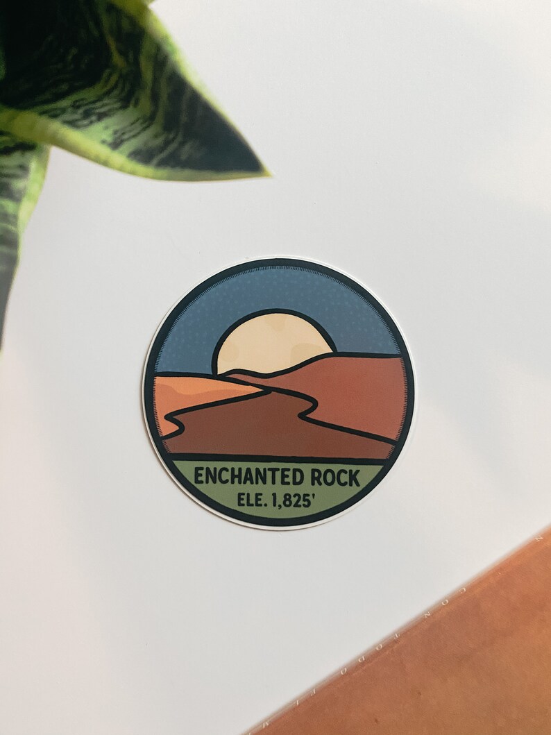 Puede incluir: Una pegatina redonda con un dise&ntilde;o de paisaje que representa una puesta de sol sobre un paisaje des&eacute;rtico. La pegatina tiene un borde azul y el texto "ENCHANTED ROCK ELE. 1,825'". La pegatina est&aacute; sobre una superficie blanca.