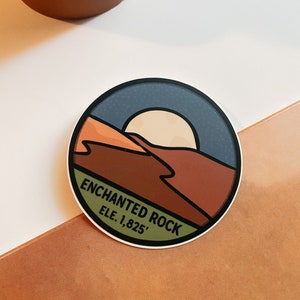 Puede incluir: Pegatina redonda con dise&ntilde;o de paisaje, sol, monta&ntilde;as y texto "ENCHANTED ROCK ELE. 1.825'". Borde blanco y cielo azul oscuro.