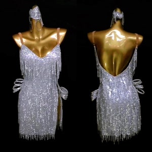 May include: A silver sequined dance dress with fringe detailing. The dress has a V-neckline and a low back. The dress is sleeveless and has a fitted silhouette.