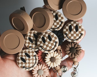 May include: A set of brown and white patterned retractable badge holders with smiley face designs. The holders are attached to brown and white patterned lanyards with brown and white patterned charms.