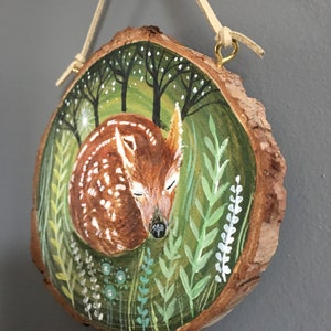 Puede incluir: Una rodaja de madera pintada a mano con un cervatillo durmiendo rodeado de follaje verde y blanco. El cervatillo es marr&oacute;n con manchas blancas y tiene los ojos cerrados. El fondo es verde oscuro con puntos blancos.