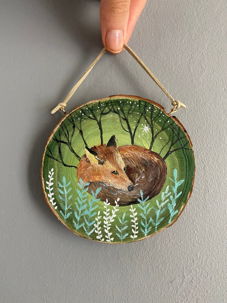 K&ouml;nnte beinhalten: Eine handbemalte Holzscheibe mit einem Fuchs, der sich in einer gr&uuml;nen Waldszene zusammengerollt hat. Der Fuchs ist braun und orange mit wei&szlig;en Akzenten. Der Wald ist in Gr&uuml;nt&ouml;nen mit wei&szlig;en Sternen und blauen Blumen bemalt.