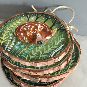 Puede incluir: Un adorno de madera pintado a mano que representa un cervatillo durmiendo rodeado de follaje verde y un cielo estrellado. El adorno est&aacute; hecho de varias rodajas de madera apiladas.