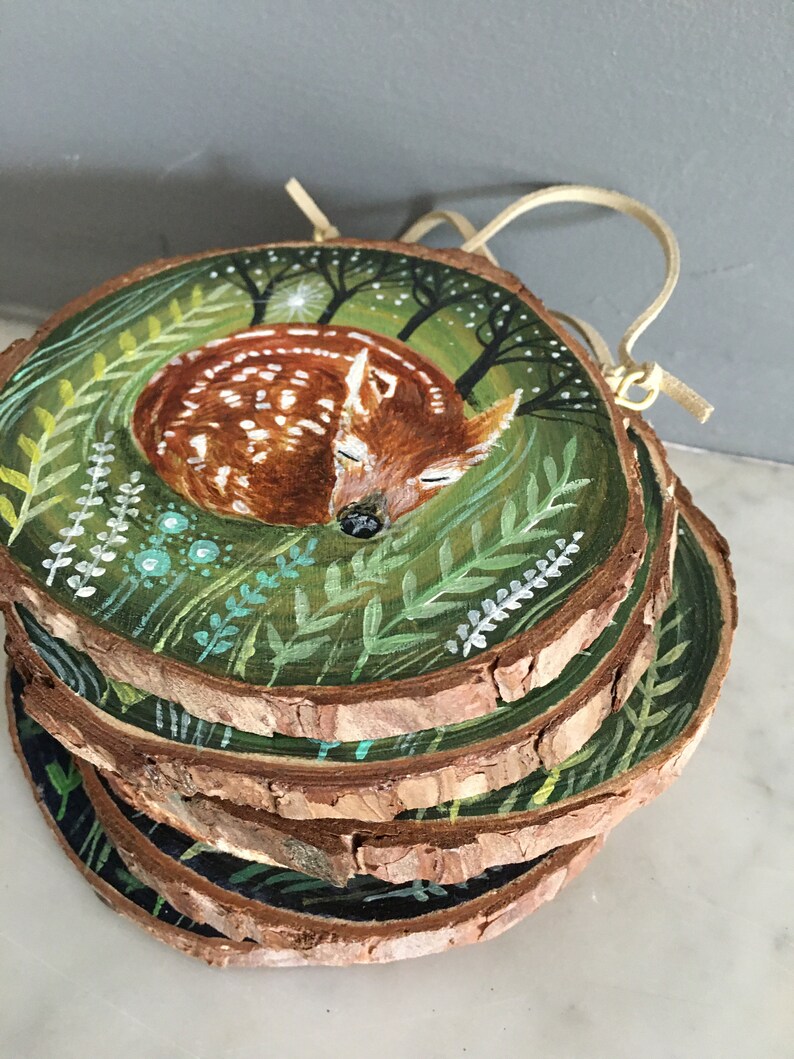 Puede incluir: Un adorno de madera pintado a mano que representa un cervatillo durmiendo rodeado de follaje verde y un cielo estrellado. El adorno est&aacute; hecho de varias rodajas de madera apiladas.