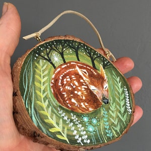 Puede incluir: Una rodaja de madera pintada a mano con un cervatillo durmiendo en un entorno de bosque. El cervatillo es marr&oacute;n con manchas blancas y est&aacute; rodeado de follaje verde y &aacute;rboles. La rodaja tiene un cord&oacute;n de cuero para colgar.