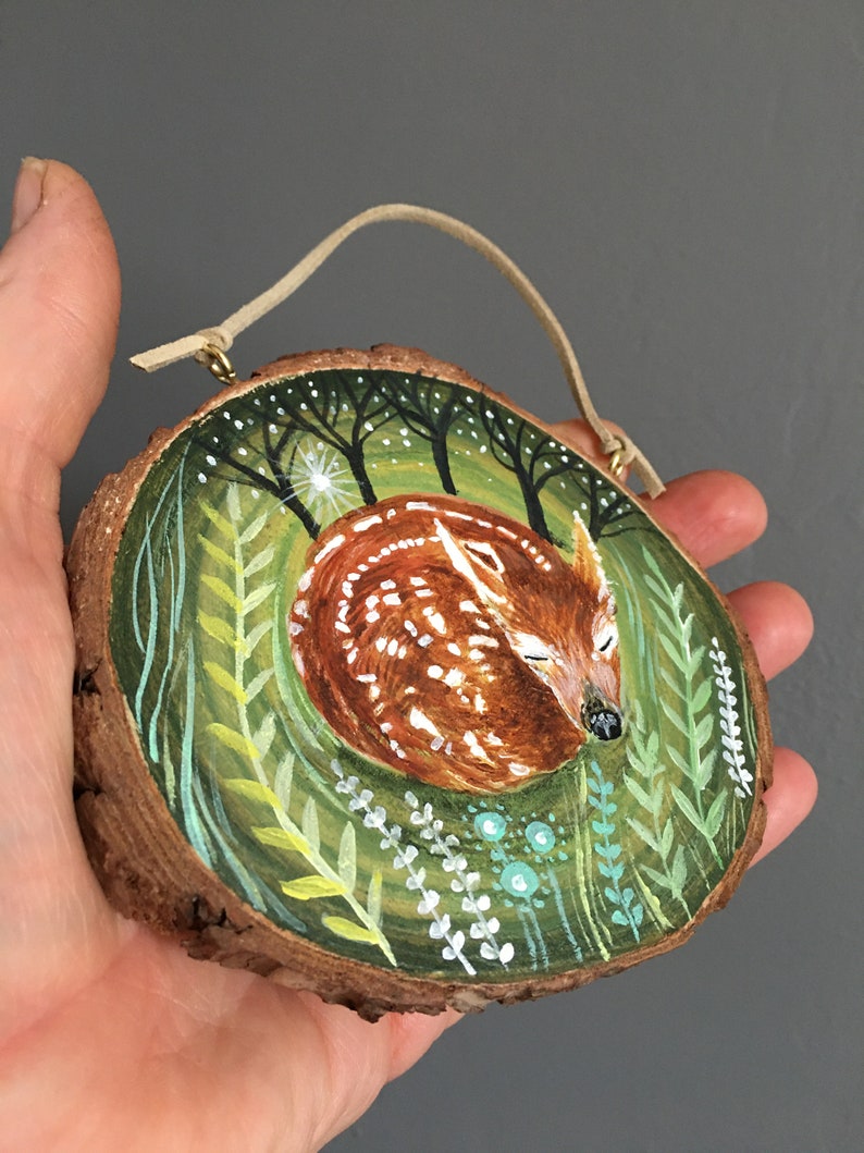 Puede incluir: Una rodaja de madera pintada a mano con un cervatillo durmiendo en un entorno de bosque. El cervatillo es marr&oacute;n con manchas blancas y est&aacute; rodeado de follaje verde y &aacute;rboles. La rodaja tiene un cord&oacute;n de cuero para colgar.