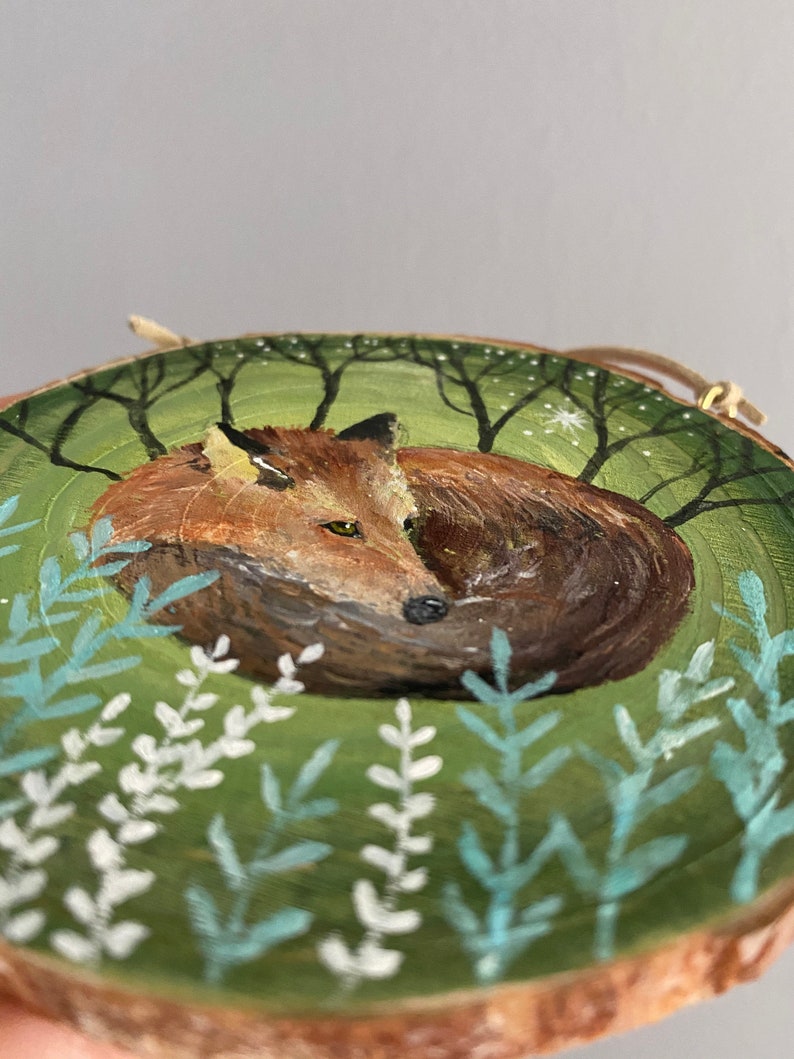 K&ouml;nnte beinhalten: Eine handbemalte Holzscheibe mit einem Fuchs, der sich in einer gr&uuml;nen Waldlandschaft zusammengerollt hat. Der Fuchs ist in Braun- und Oranget&ouml;nen gemalt, mit einem wei&szlig;en Stern im Hintergrund.