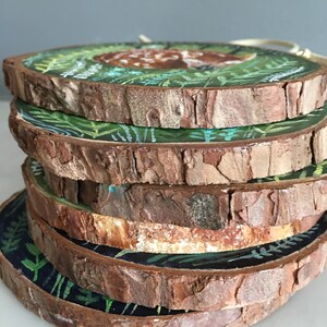 Puede incluir: Una pila de seis rodajas de madera redondas, cada una pintada con un dise&ntilde;o diferente que presenta un ciervo en un entorno de bosque. Las rodajas de madera est&aacute;n apiladas una encima de la otra, con la rodaja superior mostrando un ciervo acostado en un bosque verde.