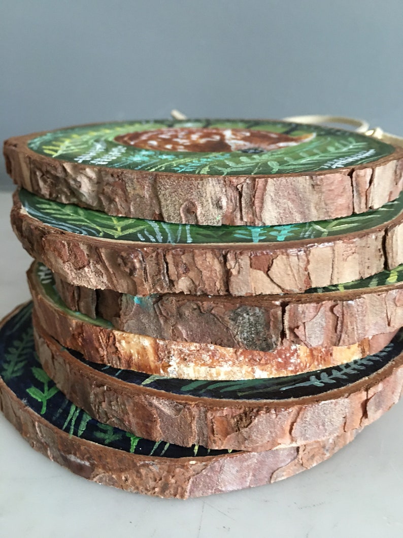 Puede incluir: Una pila de seis rodajas de madera redondas, cada una pintada con un dise&ntilde;o diferente que presenta un ciervo en un entorno de bosque. Las rodajas de madera est&aacute;n apiladas una encima de la otra, con la rodaja superior mostrando un ciervo acostado en un bosque verde.