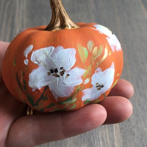 May include: A small orange pumpkin painted with white flowers and gold accents. The pumpkin is being held in a hand.