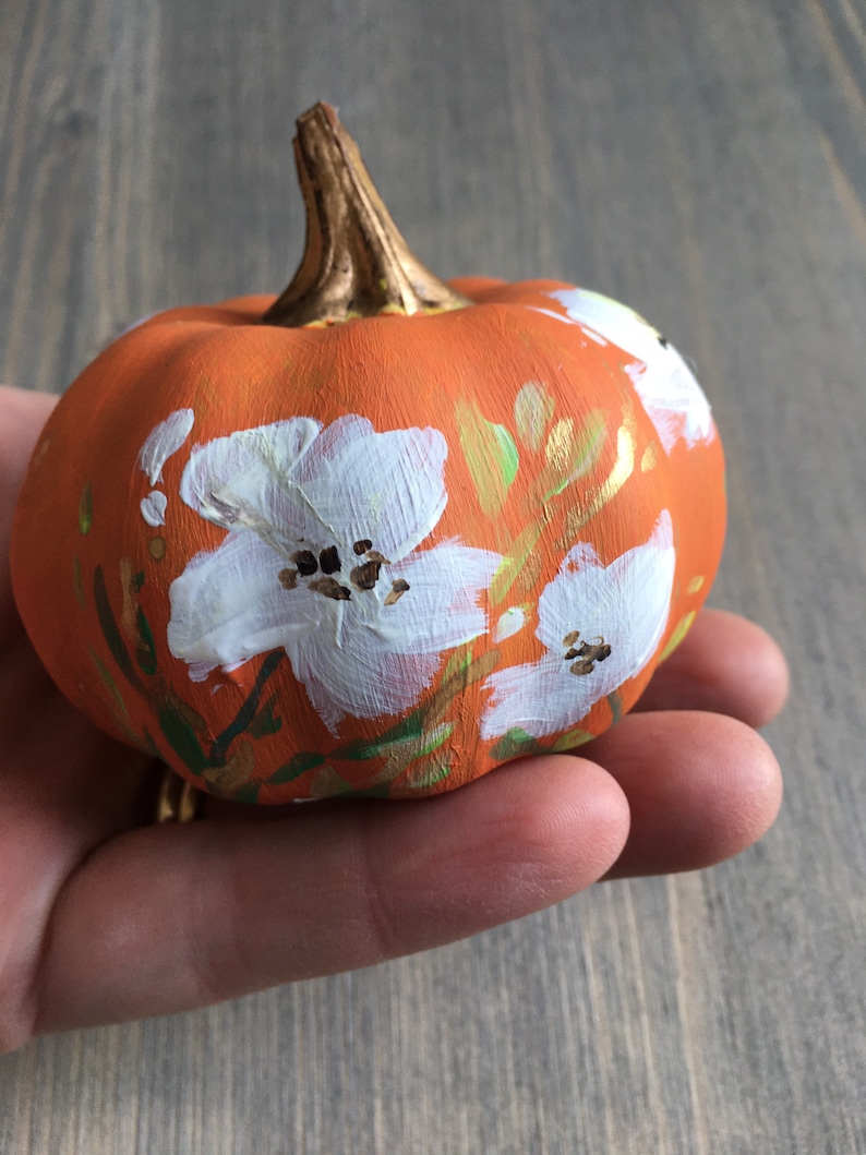 May include: A small orange pumpkin painted with white flowers and gold accents. The pumpkin is being held in a hand.