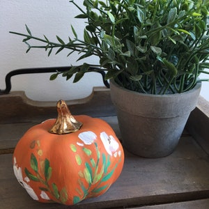 May include: A small orange pumpkin painted with white flowers and green leaves. The pumpkin is sitting on a wooden surface next to a potted green plant.