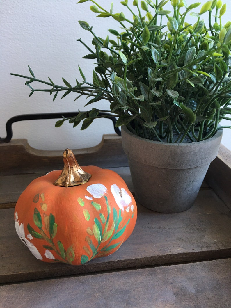 May include: A small orange pumpkin painted with white flowers and green leaves. The pumpkin is sitting on a wooden surface next to a potted green plant.