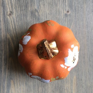 May include: A small orange pumpkin with a gold stem and white painted flowers.