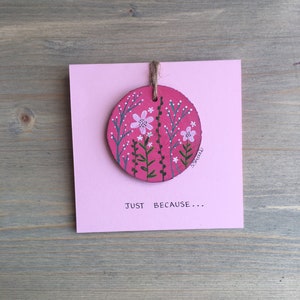 Puede incluir: Un círculo de madera rosa con un diseño floral pintado y las palabras "JUST BECAUSE..." en una tarjeta rosa.