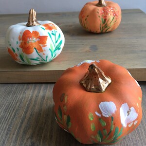 May include: Three painted pumpkins with floral designs. One pumpkin is white with orange flowers, and two pumpkins are orange with white and green flowers.
