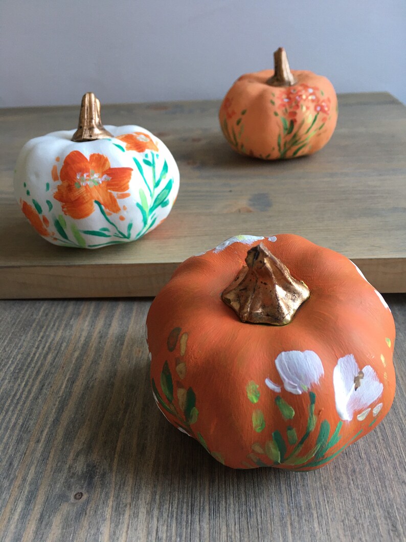 May include: Three painted pumpkins with floral designs. One pumpkin is white with orange flowers, and two pumpkins are orange with white and green flowers.