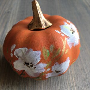 May include: A small orange pumpkin with a gold stem is painted with white flowers and green leaves.