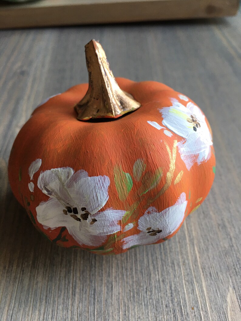 May include: A small orange pumpkin with a gold stem is painted with white flowers and green leaves.