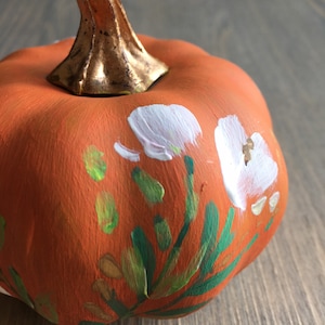 May include: A small orange pumpkin painted with white flowers, green leaves, and gold accents.