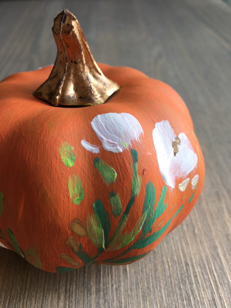 May include: A small orange pumpkin painted with white flowers, green leaves, and gold accents.