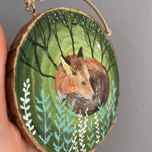 K&ouml;nnte beinhalten: Ein handbemaltes Holz-Ornament mit einem Fuchs, der sich in einer gr&uuml;nen Waldszene mit wei&szlig;en Blumen zusammengerollt hat. Das Ornament ist rund und hat eine Schnur zum Aufh&auml;ngen.