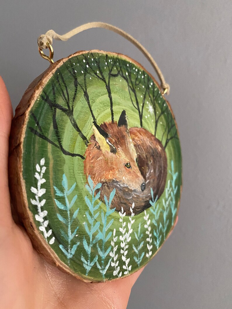 K&ouml;nnte beinhalten: Ein handbemaltes Holz-Ornament mit einem Fuchs, der sich in einer gr&uuml;nen Waldszene mit wei&szlig;en Blumen zusammengerollt hat. Das Ornament ist rund und hat eine Schnur zum Aufh&auml;ngen.