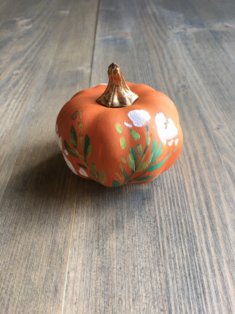 May include: A small orange pumpkin painted with white flowers and green leaves. The pumpkin has a gold stem.