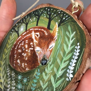Puede incluir: Un adorno de madera pintado a mano que representa un cervatillo durmiendo en un entorno de bosque. El cervatillo es marr&oacute;n con manchas blancas y est&aacute; rodeado de follaje verde y flores blancas. El adorno est&aacute; sujeto a un cord&oacute;n de cuero marr&oacute;n con un lazo de metal dorado.