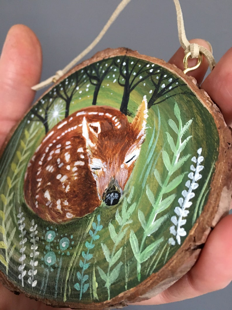 Puede incluir: Un adorno de madera pintado a mano que representa un cervatillo durmiendo en un entorno de bosque. El cervatillo es marr&oacute;n con manchas blancas y est&aacute; rodeado de follaje verde y flores blancas. El adorno est&aacute; sujeto a un cord&oacute;n de cuero marr&oacute;n con un lazo de metal dorado.