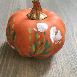 May include: A small orange pumpkin with a gold stem is painted with white flowers and green leaves.