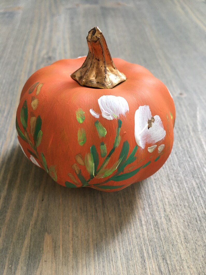 May include: A small orange pumpkin with a gold stem is painted with white flowers and green leaves.