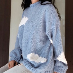 Puede incluir: Un suéter de punto azul claro con aplicaciones de nubes blancas. El suéter tiene cuello redondo y mangas largas.