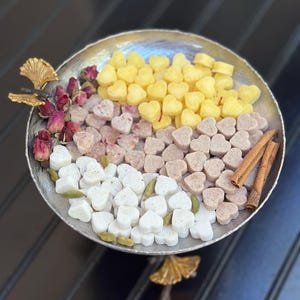May include: A silver serving dish with a gold leaf accent holds heart-shaped treats in yellow, pink, white, and brown. Dried rose buds, green cardamom pods, and cinnamon sticks add to the presentation. The dish is on a dark surface.