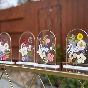 May include: Four clear acrylic table number signs with dried flowers and white script. Each sign has a copper-colored edge and a white base. The numbers are Five, Six, Seven, and Eight. The flowers are pink, white, yellow, and purple.