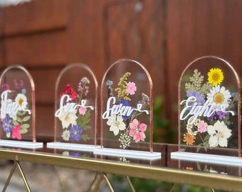 May include: Four clear acrylic table number signs with dried flowers and white script. Each sign has a copper-colored edge and a white base. The numbers are Five, Six, Seven, and Eight. The flowers are pink, white, yellow, and purple.