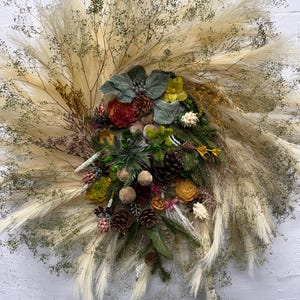 May include: A decorative wreath featuring a large circular arrangement of cream-colored pampas grass and dried baby's breath. The center is adorned with pinecones, green foliage, yellow and red flowers, and small brown seed pods.