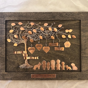 May include: A rectangular wooden frame containing a wire tree sculpture with copper-colored leaves. Hanging from the branches are copper shapes engraved with the names Olivia, Luke, Ella, Oscar, Frank, and Leo. A copper plaque reads October 15, 2019.